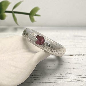 May include: A silver ring with a textured, matte finish featuring a single, small, round, deep pink gemstone embedded in the centre of the band. The metal surface has a rough, organic appearance that contrasts with the smooth, polished stone.