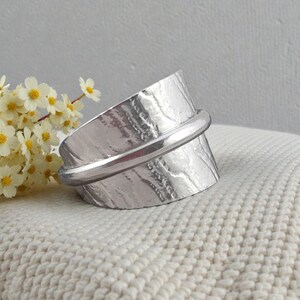 May include: A silver-colored ring with a textured surface and a smooth band. The ring is set against a white, textured background, with a sprig of small white flowers with yellow centers in the background.