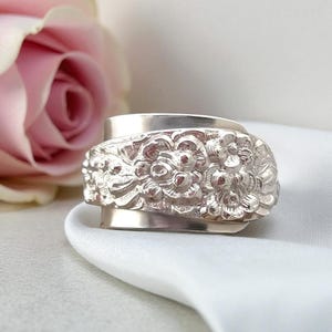 May include: A silver spoon ring featuring a wide band with a detailed embossed floral pattern. The metal surface is polished and reflective, showcasing intricate sculpted flower petals and leaves that wrap around the circumference of the band.