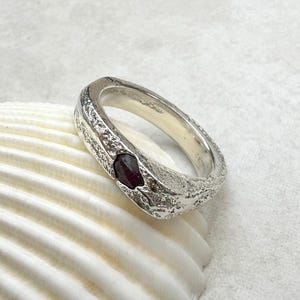 May include: A silver ring with a textured band and a dark red gemstone. The ring is set on a white seashell, showcasing its design and color. The gemstone is oval-shaped and adds a pop of color to the ring.