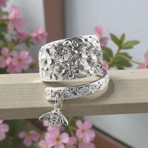May include: A silver-colored spoon ring with floral detailing and a bee charm. The ring is wrapped around a wooden surface, with pink flowers in the background. The ring's design features embossed flowers and a dangling bee.