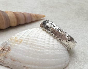 May include: A textured silver ring with an irregular surface, resting on a white seashell. The ring's design has a hammered or organic appearance. A brown and tan seashell is in the background.