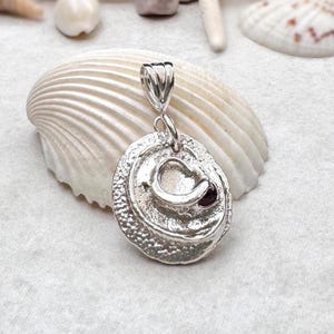 May include: A silver pendant featuring a textured, circular base with a raised, irregular center and a small, dark red gemstone set within the metal. The pendant includes a polished silver bail at the top for attachment to a chain or cord.