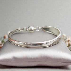 May include: A silver spoon bracelet with a floral design, displayed on a satin cushion. The bracelet has a ball clasp and is set against a grey backdrop with small white flowers. The bracelet's length is approximately 17 cm.
