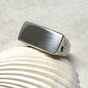 May include: A silver ring with a rectangular face. The ring has a textured band and a smooth, polished top. The ring is displayed on a white seashell, creating a contrast between the textures and colors.