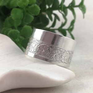 May include: A silver ring featuring a floral design. The wide band of the ring is decorated with an embossed pattern of flowers and leaves. The ring is shown on a white surface, with green foliage in the background.