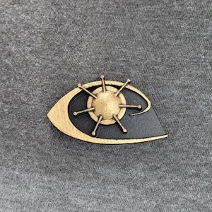 以下が含まれることがあります： 中央に太陽のようなデザインが施された、金と黒の目の形をしたピン。目の形は金の縁取りで、内側は黒。太陽は、放射状のスポークを持つ金色の中心部を持っています。