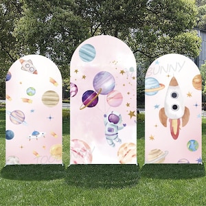 Może przedstawiać: Trzy łukowate panele z motywem kosmicznym. Każdy panel ma jasnoróżowe tło z planetami, gwiazdami i rakietami. Jeden panel ma u góry napis "BUNNY". Astronauta unosi się z balonami w kształcie planet.