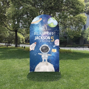 May include: A blue and white birthday party decoration with an astronaut theme. The sign reads "3, 2, 1... BLAST OFF! JACKSON is turning 5." The design includes planets, a rocket ship, and an astronaut standing on a moon.