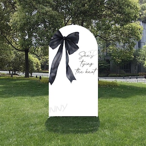 May include: A white wedding sign with a black bow and the text "She's tying the knot." The sign is arched at the top and stands on a grassy lawn, with trees and a building in the background.
