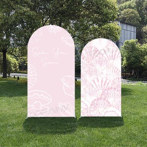 May include: Two arched pink signs with a seashell theme. One sign has the text "Sea You Soon" and seashell illustrations. The other sign features a pattern of white seashells. Both signs are on a green base.