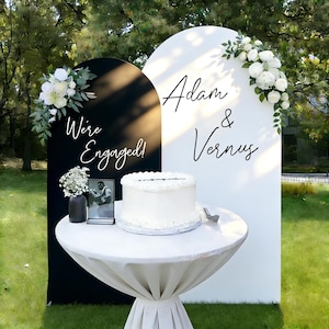 Puede incluir: Montaje para una celebración de compromiso al aire libre con una tarta blanca, una foto y dos paneles arqueados. Un panel es negro con el texto "We're Engaged!" y el otro es blanco con los nombres "Adam & Vernus". Arreglos florales adornan los paneles.