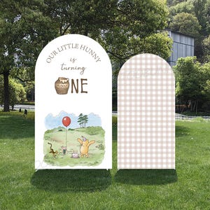May include: Two arched signs for a first birthday celebration. One sign features Winnie the Pooh characters with the text "Our Little Hunny is turning ONE." The other sign has a tan and white checkered pattern.