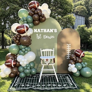 May include: A football-themed party backdrop with sage green and brown arched panels, a balloon garland in white, brown, and sage, and football-shaped balloons. A white high chair sits on a green rug printed like a football field with text.