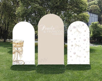May include: Three arched wedding backdrops on a grassy lawn. One backdrop features a champagne glass illustration with a bow and pearls. The center backdrop has the text "Pearls & Prosecco". The third backdrop has a pearl and bow pattern.