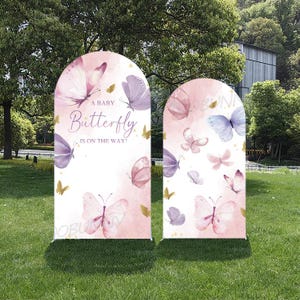 Op de afbeelding: Twee boogvormige, roze en witte achtergrondbanners met aquarel vlinders in roze, paarse en blauwe tinten. De tekst "A Baby Butterfly Is On The Way!" staat op één banner. De banners staan op een grasveld.