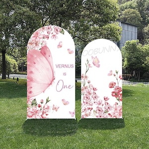 Può includere: Due cartelli bianchi a forma di arco con disegni floreali e farfalle rosa. Un cartello presenta una grande farfalla rosa e il testo "VERNUS IS One". L'altro cartello ha una composizione floreale e il testo "OOBUNN".