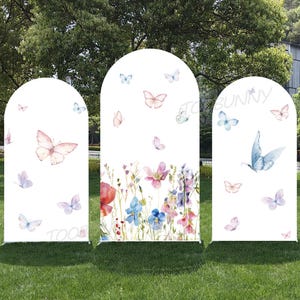 Può includere: Tre pannelli bianchi ad arco decorati con farfalle e fiori ad acquerello. I pannelli sono posizionati su erba verde, con uno sfondo naturale di alberi e cielo. I pannelli hanno il testo "TOO BUNNY".