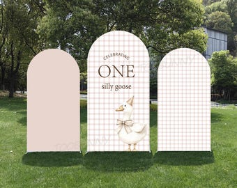 May include: Three arched panels set on grass. The center panel features a goose illustration with a party hat and bow, and the text "Celebrating ONE silly goose". The panels have a pink and white plaid pattern.