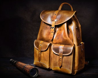 May include: A brown leather backpack with two large front pockets and a buckle closure. The backpack is sitting on a wooden surface with a vintage telescope.