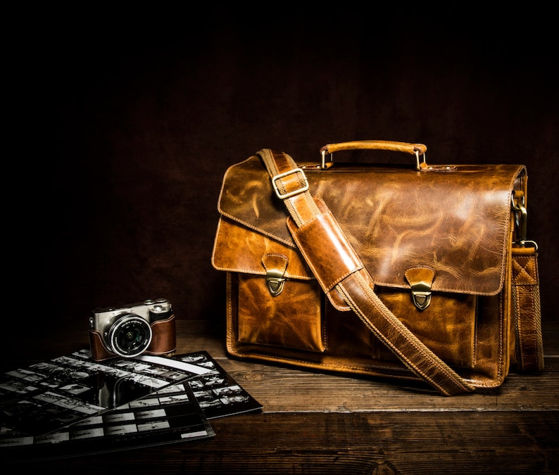 May include: A brown leather briefcase with a long strap and a silver buckle. The briefcase has two compartments and a flap closure. The briefcase is sitting on a wooden surface next to a vintage camera.