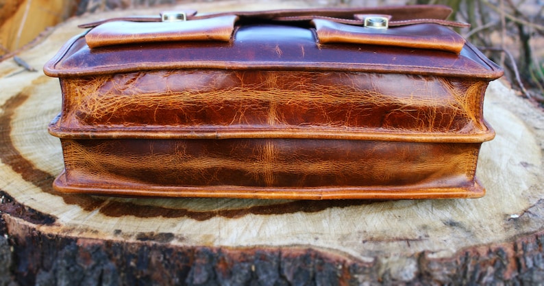 May include: A brown leather briefcase with two front pockets and a flap closure. The briefcase has a distressed look and is made of high-quality leather.