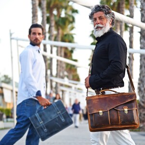 May include: Two men walking down a sidewalk, one carrying a brown leather briefcase with a strap, the other carrying a blue briefcase.