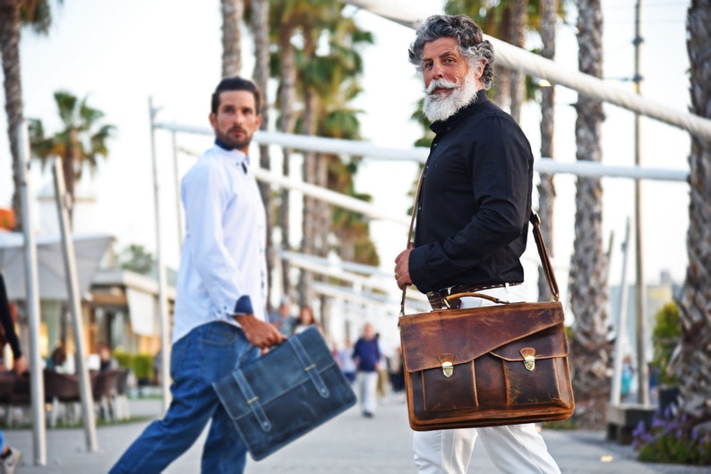 May include: Two men walking down a sidewalk, one carrying a brown leather briefcase with a strap, the other carrying a blue briefcase.