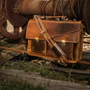 May include: A brown leather briefcase with a long strap and two front pockets. The bag is hanging from a rusty metal structure.