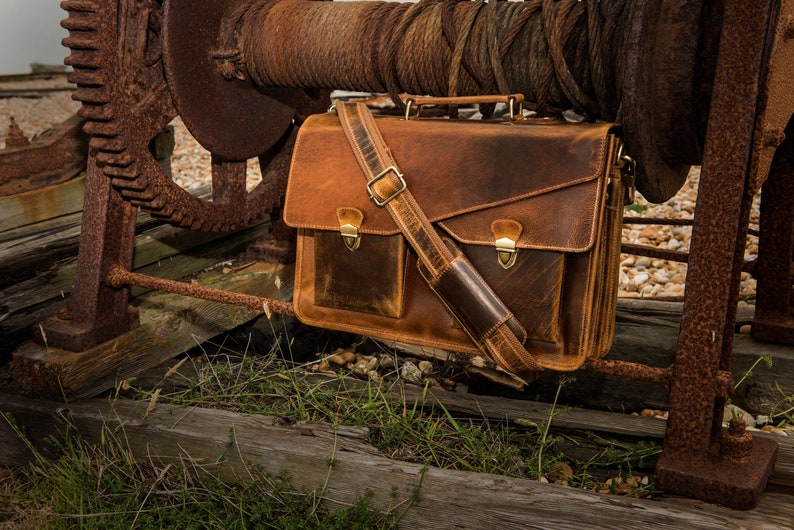 May include: A brown leather briefcase with a long strap and two front pockets. The bag is hanging from a rusty metal structure.