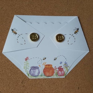 May include: A light blue, folded paper card with a scalloped edge, featuring illustrations of bees and honey pots. Two gold buttons secure the card. The card's design includes a whimsical bee flight path and colorful floral accents.
