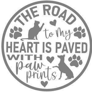 Pode incluir: Um círculo cinza com o texto "The road to my heart is paved with paw prints" em fonte branca. O círculo também inclui um gato, um cão, pegadas e corações.