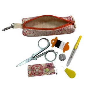 May include: A fabric pouch with a pink and orange circular pattern and an orange zipper, accompanied by metal scissors, a spool with black and white thread, a silver thimble, a yellow seam ripper, a yellow needle threader, and a small needle case.