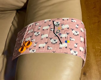 May include: A pink fabric armrest organizer with a sheep print. It has a pocket and is filled with sewing supplies, including scissors, pins, and thread.