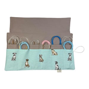 May include: A light blue fabric roll with a pattern of dogs. The roll has six pockets that hold various coloured knitting needles. The roll is closed with a white strap.