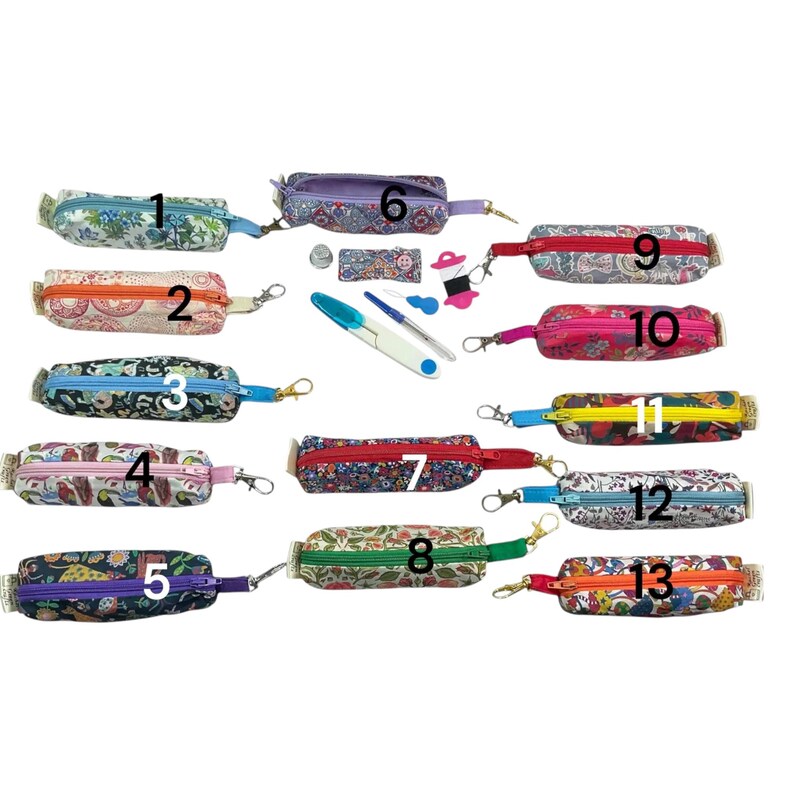 May include: Thirteen small, rectangular fabric zipper pouches with numbered labels, each featuring distinct floral or animal patterns and a metal lobster clasp. Included are sewing accessories: a thimble, a seam ripper, and a thread spool.
