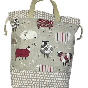 May include: A beige canvas tote bag with a drawstring closure. The bag features a white polka dot pattern on the bottom and a print of sheep with different coloured patterns on the top. The bag has two beige handles.