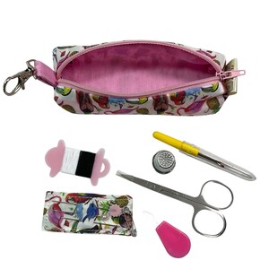 May include: A small zippered pouch with a tropical bird and pineapple print, featuring a pink interior and a metal clip. Included are sewing accessories: a spool of thread, a metal thimble, a yellow seam ripper, silver scissors, and a needle case.