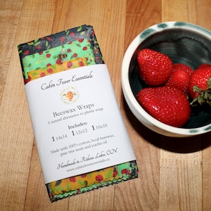 May include: A set of three beeswax food wraps with a bee and strawberry design. The wraps are made with 100% cotton, beeswax, pine tree resin, and jojoba oil. The wraps are handmade in Rideau Lakes, ON. The text on the label reads "Cabin Fever Essentials Beeswax Wraps A natural alternative to plastic wrap Includes: 1 14x14 1 12x12 1 10x10 Made with 100% cotton, local beeswax, pine tree resin and jojoba oil Handmade in Rideau Lakes, ON www.cabinfeveressentials.ca"