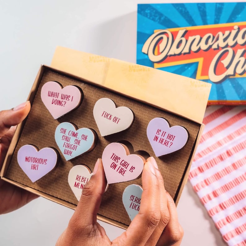 Hands holding a box of Bagstock & Bumble Menopause Obnoxious Chocs, filled with pastel-topped Belgian chocolate hearts and brutally honest messages.