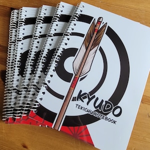 Puede incluir: Una pila de cuadernos con espiral y una cubierta blanca que muestra un blanco de tiro con arco en blanco y negro con una flecha roja y marrón. El texto "KYUDO TEKICHUINOTEBOOK" está impreso en la cubierta.