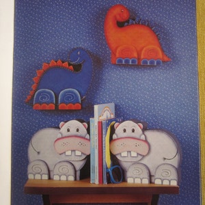 May include: Two wooden bookends shaped like hippos, painted in shades of gray, with pink noses and white teeth. The bookends are holding two books, one with a rainbow design and the other with a blue and white design.  Two wooden dinosaur bookends are on the wall behind the hippos. One dinosaur is blue with red feet and the other is orange with blue feet.