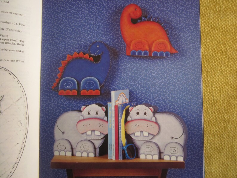 May include: Two wooden bookends shaped like hippos, painted in shades of gray, with pink noses and white teeth. The bookends are holding two books, one with a rainbow design and the other with a blue and white design.  Two wooden dinosaur bookends are on the wall behind the hippos. One dinosaur is blue with red feet and the other is orange with blue feet.