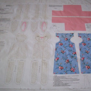 May include: A fabric pattern for sewing a bunny doll. The pattern includes pieces for the body, head, ears, legs, and a dress. The fabric is white with a light brown pattern for the bunny and blue with a floral pattern for the dress.