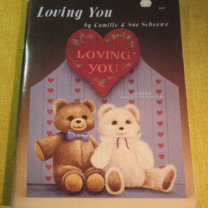 May include: A book titled "Loving You" by Camille & Sue Scheewe, featuring a painted image of two teddy bears sitting together in front of a heart-shaped sign that reads "Loving You".