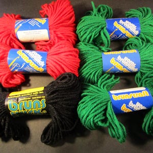 Può includere: Un assortimento di gomitoli di filato rosso, verde e nero. Ogni gomitolo è avvolto con un'etichetta che recita "Brunscraft". Il filato è disposto su una superficie scura, mostrando i colori vivaci e le trame del materiale artigianale.