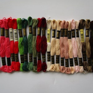 May include: Assortment of 24 embroidery floss skeins in various colors, including red, green, brown, and beige. Each skein has a black label with gold lettering and a number.