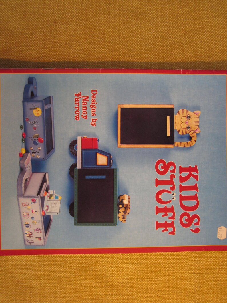 May include: A book cover with the title "Kids' Stuff" in red and white letters. The cover features illustrations of wooden toys, including a truck, a mailbox, and a toy with a chalkboard. The book is by Nancy Farrow.