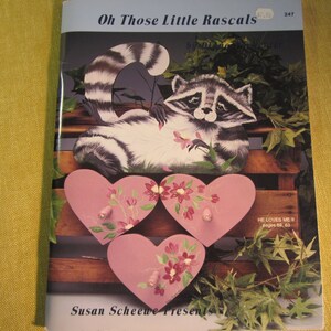 May include: A book cover with a raccoon illustration and the title "Oh Those Little Rascals". The cover also features three pink heart-shaped wooden ornaments with floral designs and the text "He Loves Me If" and "pages 62, 63".