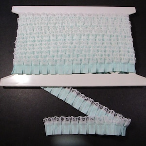 May include: A roll of ruffled lace trim in a soft, pale blue color. The lace is gathered and has a delicate, textured appearance. The trim is on a white plastic spool, with a section of the trim unrolled, showing a scalloped edge with black detailing.